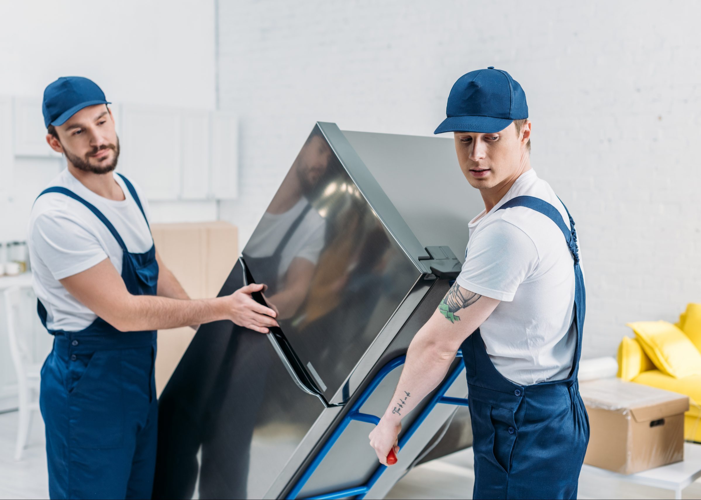 two movers in uniform using hand truck while transporting refrigerator in apartment